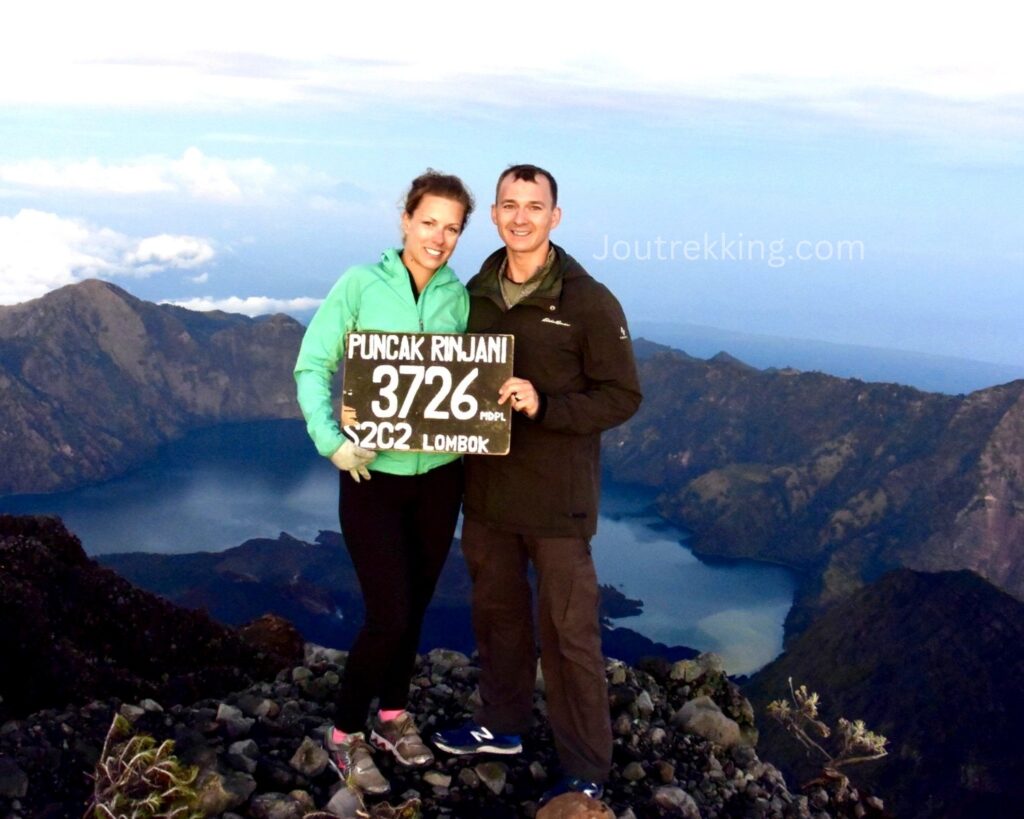 Two trekkers standing at the summit of Mount Rinjani (3,726 m) holding a summit sign, with the stunning Segara Anak Crater Lake and volcanic landscape in the background during sunrise.