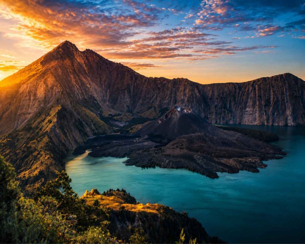 Panoramic sunrise view of Mount Rinjani's Segara Anak Crater Lake and Barujari Volcano from the Senaru Crater Rim, with dramatic cliffs, turquoise water, and golden morning light illuminating the landscape.
